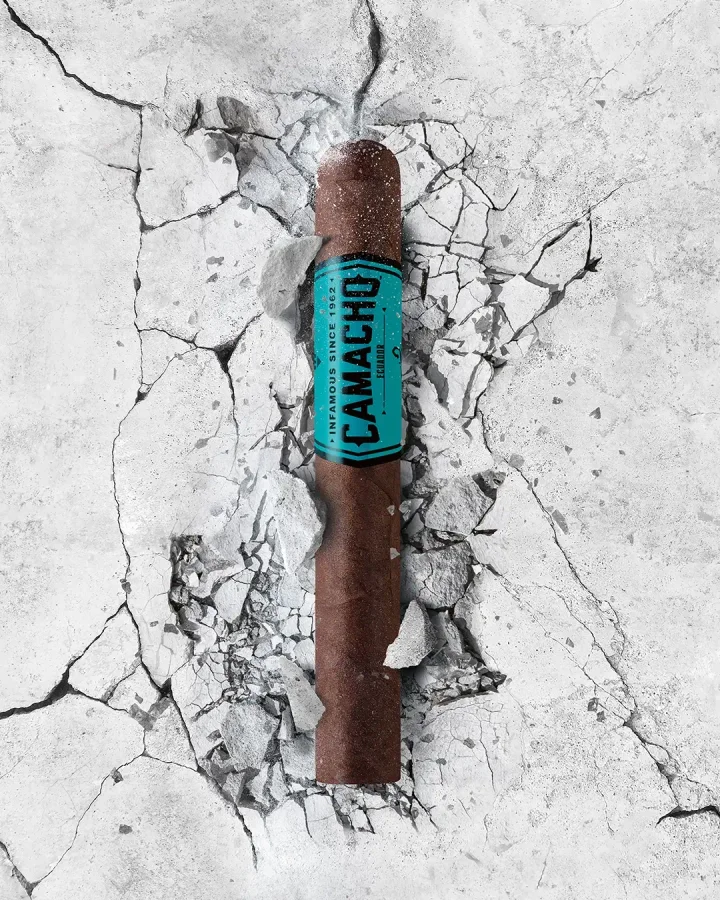 A Camacho cigar stands upright amidst a stylized illustration of a scorpion and dynamic splashes of color against a blue background.