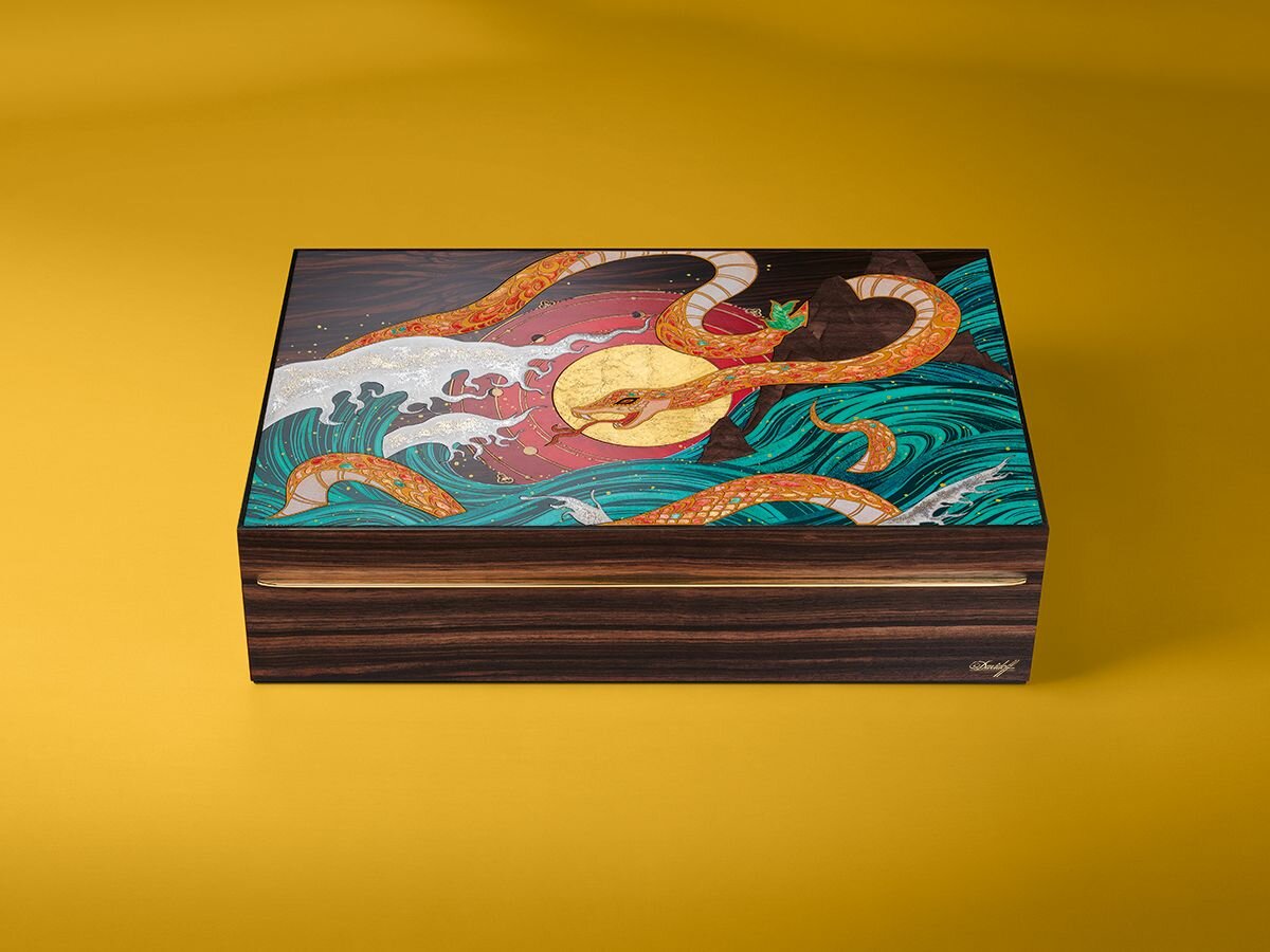 A beautifully designed "Year of the Snake" hunidor featuring swirling waves, a golden sun, and intricate serpent motifs on a vibrant yellow background.