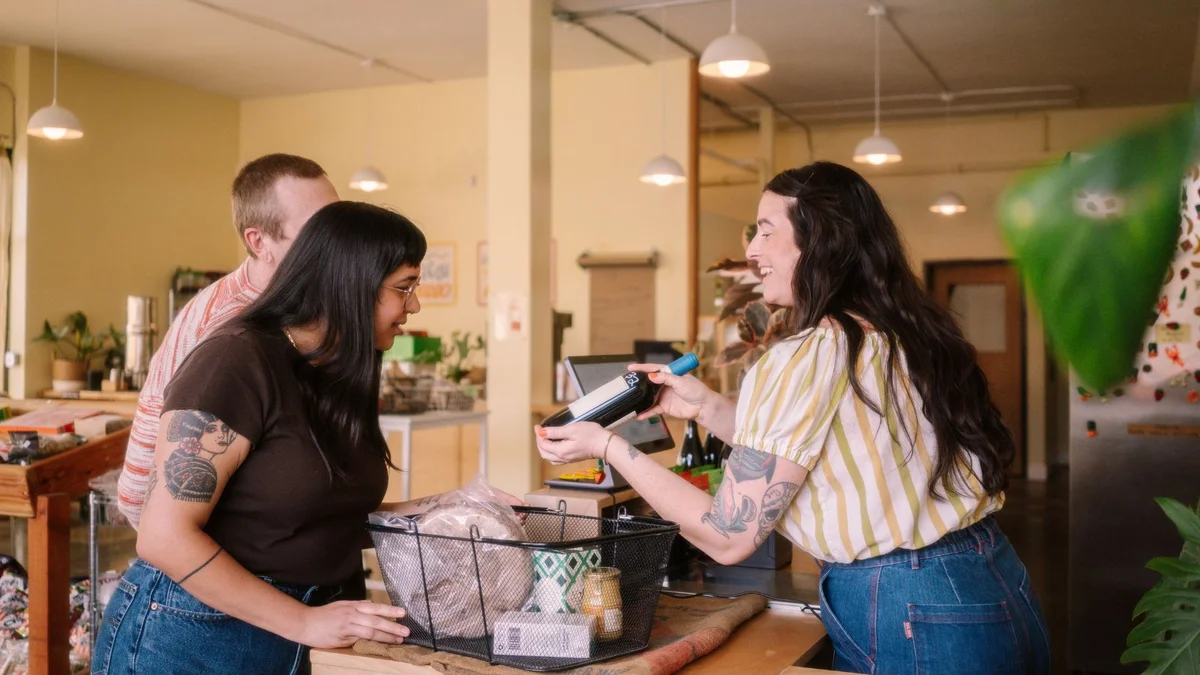 Smiling shop owner at the counter of a small grocery store, showing a bottle of wine to two customers with a basket of bread and pantry items