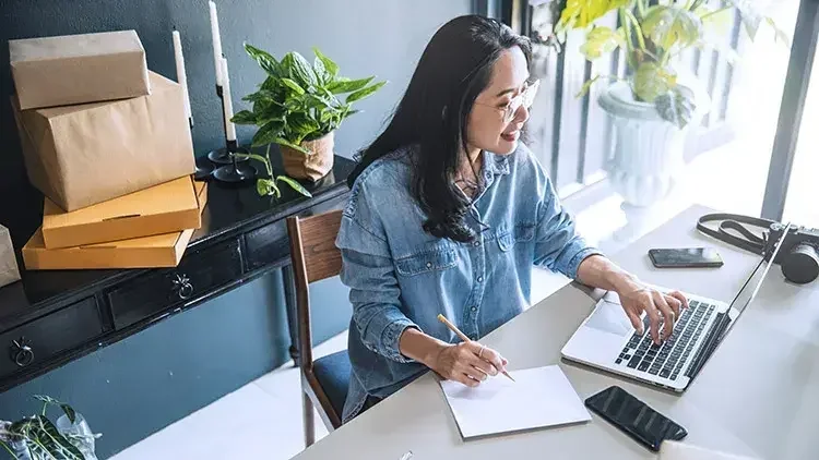 Woman smiling while sitting at a desk, looking at a laptop screen and taking notes in a notebook.
