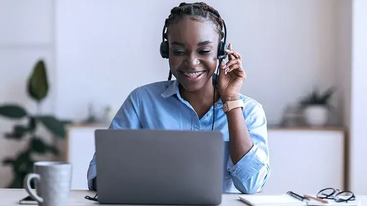 Woman wearing over-ear headphones sits and smiles at her laptop