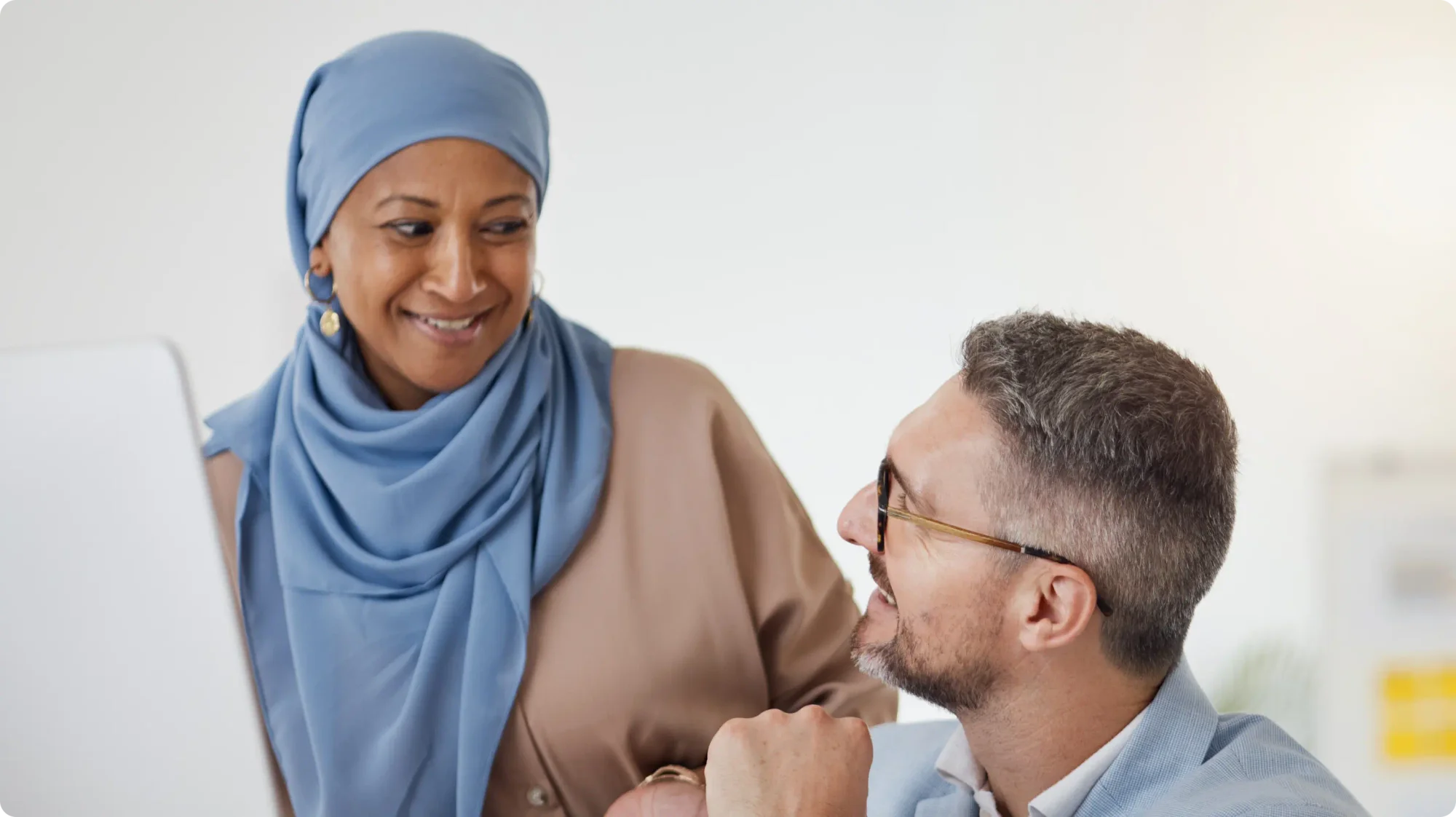 A woman in a blue hijab stands and speaks with a man wearing a business suit and glasses.