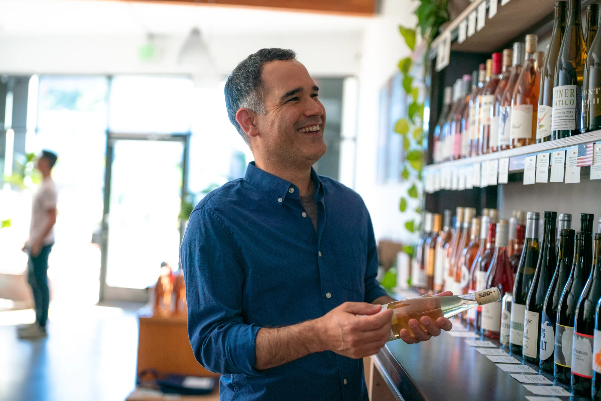 Smiling man in denim shirt holding a bottle of wine laughing off into the distance.