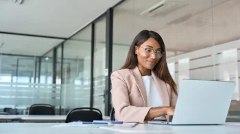 Woman smiling while working on a laptop in a bright office