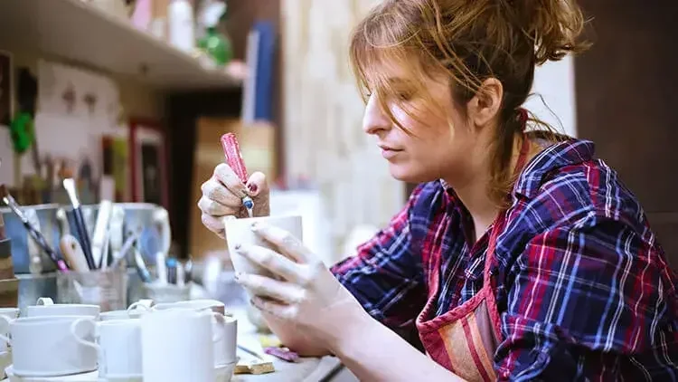 An image of a woman wearing an apron, carefully painting coffee mugs in her living room.