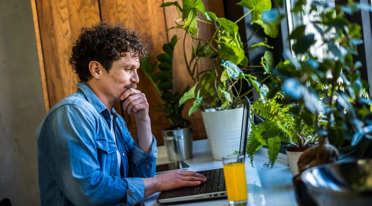 Man in denim shirt using a laptop surrounded by plants