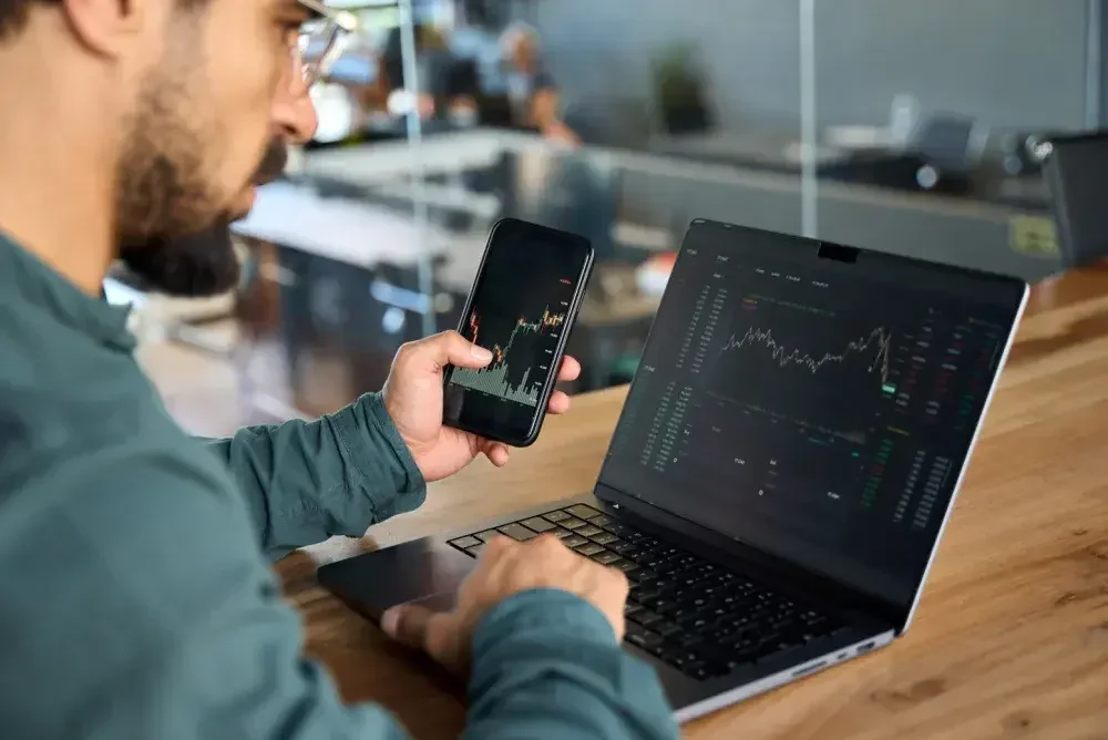 A small business owner checks the stock market on his laptop and his cell phone while he sits at a conference room table.