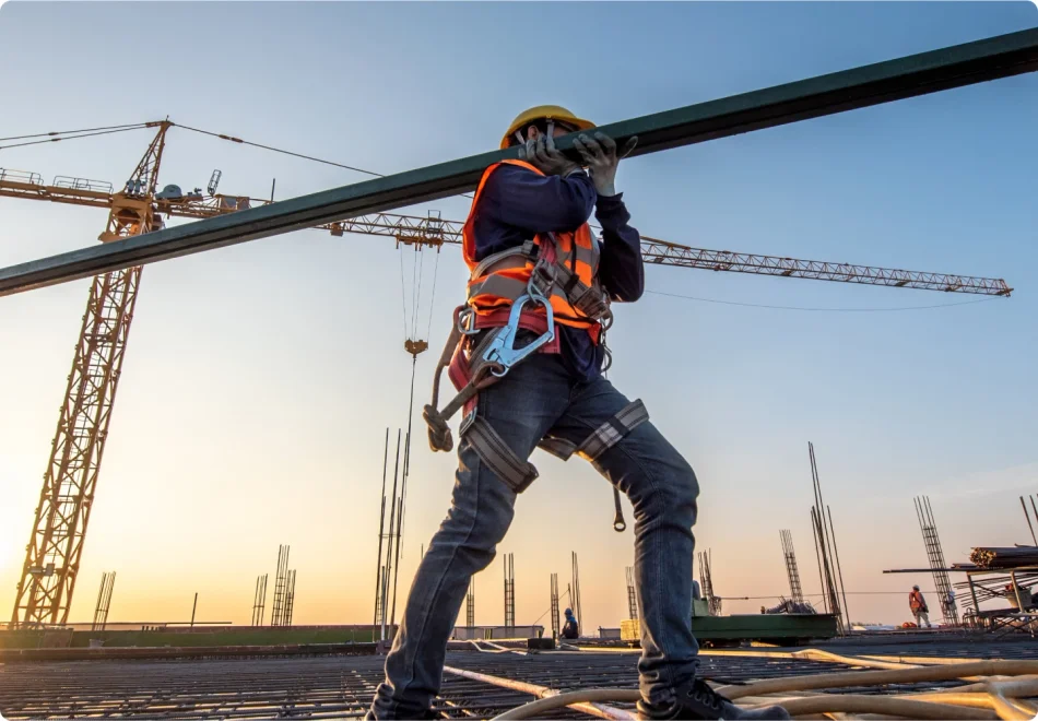 A construction worker in a high visibility vest carries a metal beam on his shoulder, through an jobsite.