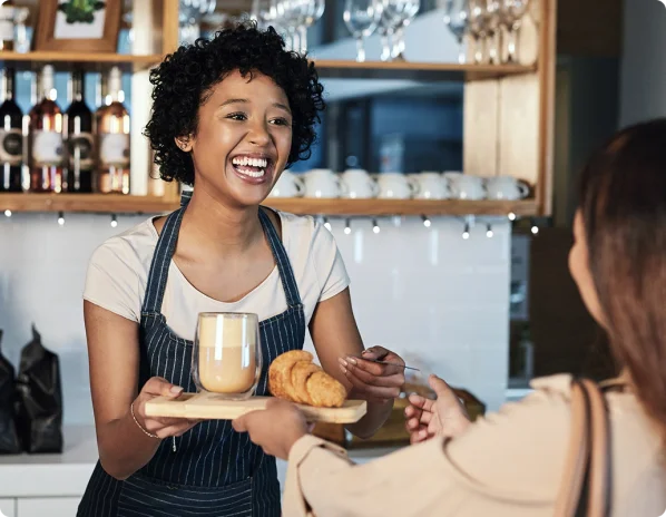 Black woman with curly hair wearing a white t-shirt and a blue and white pinstriped apron working in a café and  smiling because she got her business license from LegalZoom as she hands a woman with brown hair wearing a tan shirt her order.