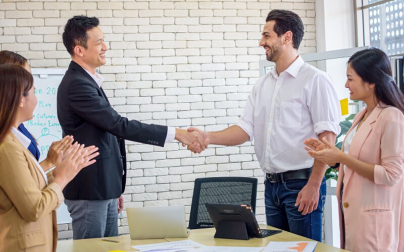 This is an image of 5 employees in a meeting, 2 of whom are shaking hands, and the rest are clapping.