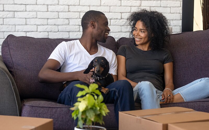 An image of a happy couple and a dog, sitting on a sofa, surrounded by boxes.