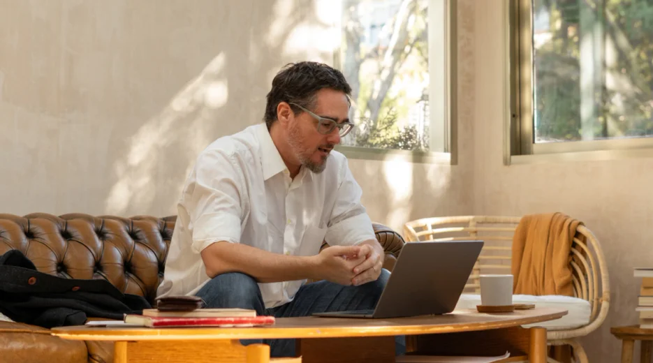 A man in casual clothes sits on a brown leather couch and reads about emergency power of attorney on his laptop.