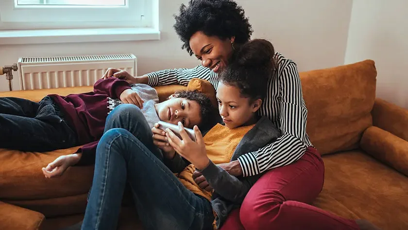 A woman hugs her children as they gather on their couch to watch a video on a smart phone.