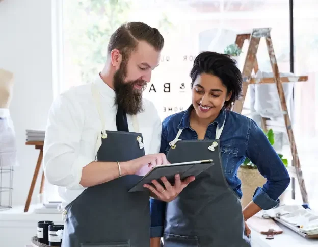 Woman with dark hair wearing a denim shirt and a grey apron looking at a LegalZoom LLC on a tablet with her business partner, a man with a beard wearing a white shirt and grey apron.