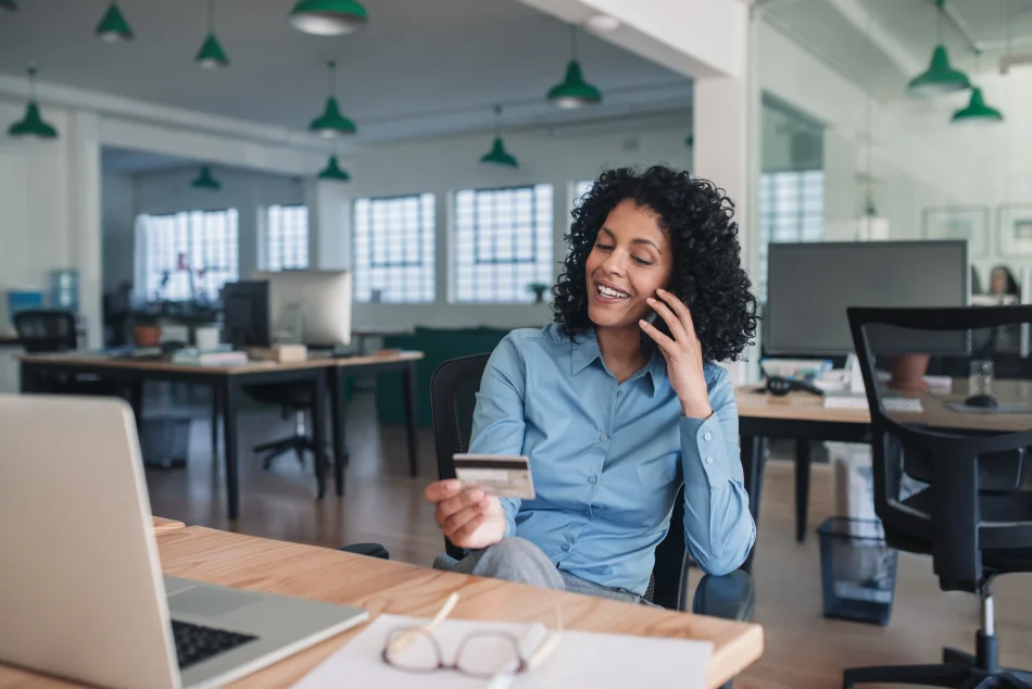 Smiling young businesswoman using her credit card to make an order on her cellphone while sitting at her desk in a large modern office