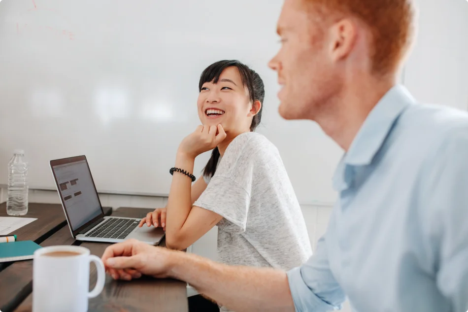 An asian woman in the background sits at her computer and laughs with a man in the foreground.