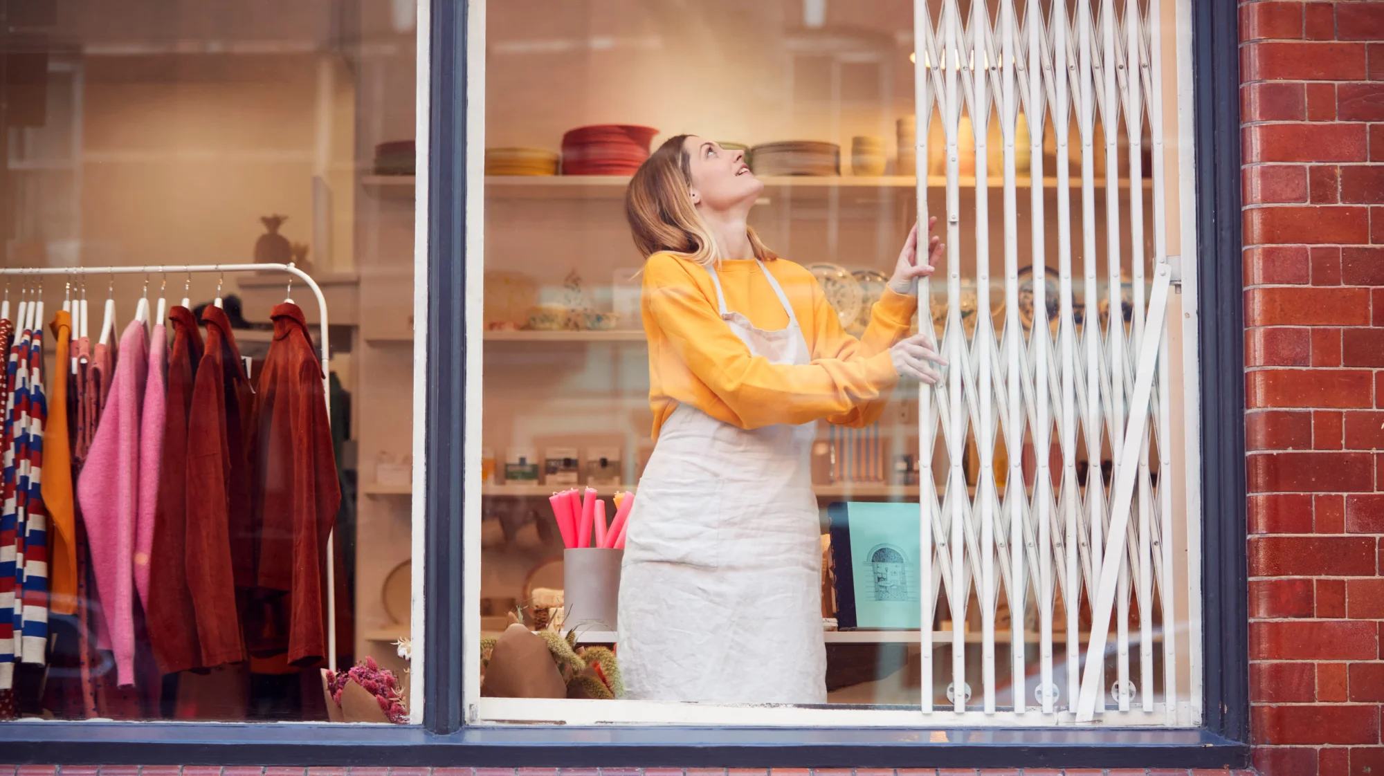 White woman storefront owner with straight light blonde hair wearing yellow shirt with a white apron smiling opens up the windows of a clothing shop