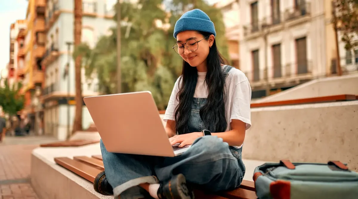 A woman wearing a beanie, glasses, and overalls sits on a bench outside with her laptop computer and reads instructions for doing a business search in Louisiana.