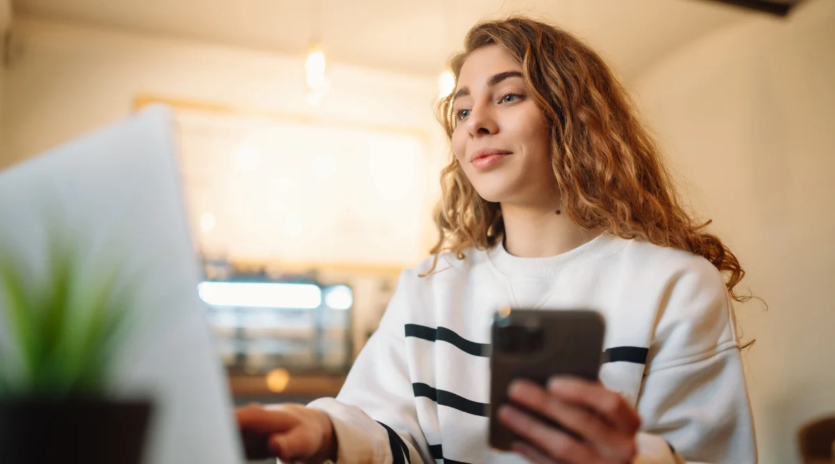 A young woman explores Hawaii business information on both her phone and computer at a crowded cafe