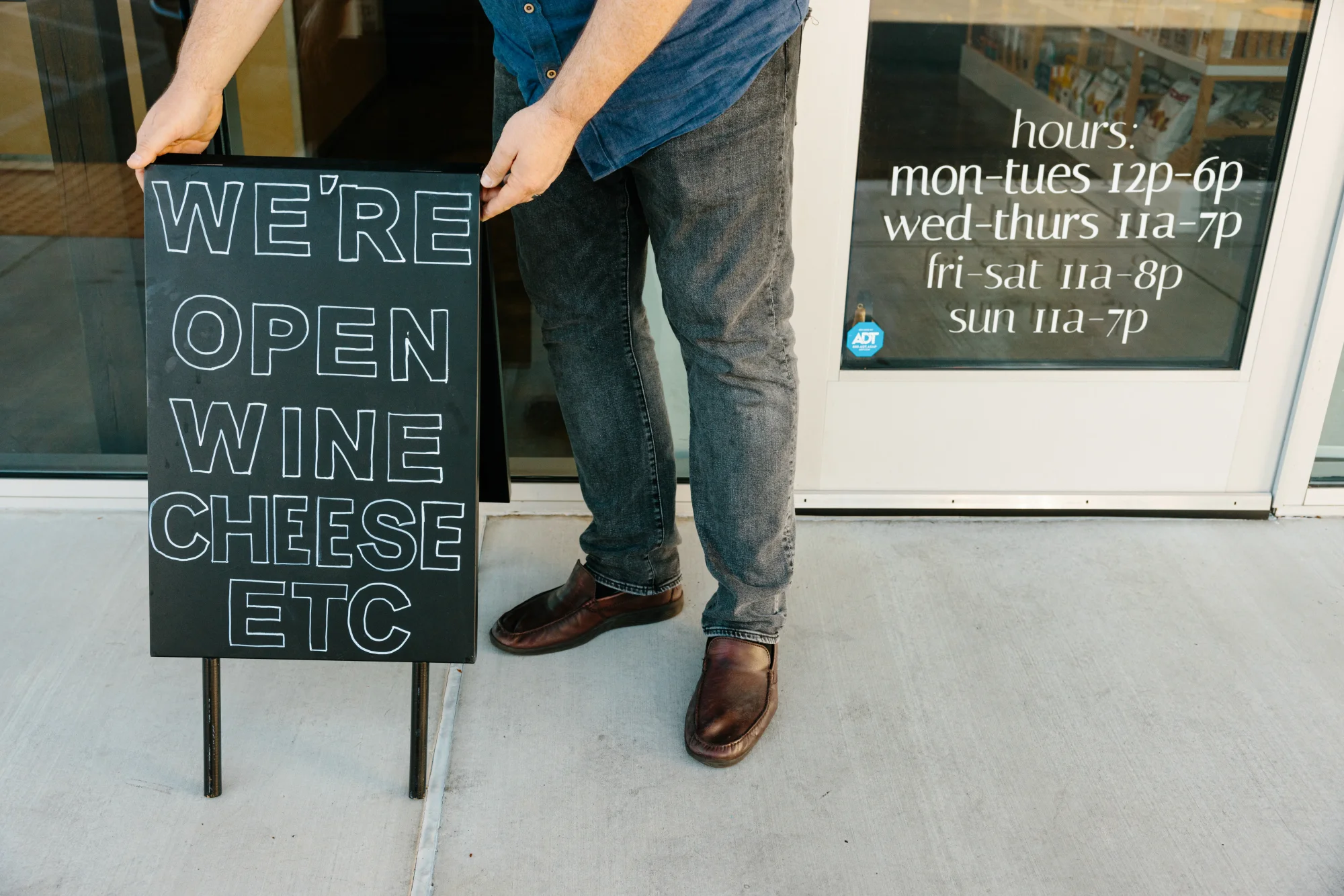 Person in black jeans setting up a sign outside of their storefront that reads "We're open! Wine, cheese, etc"