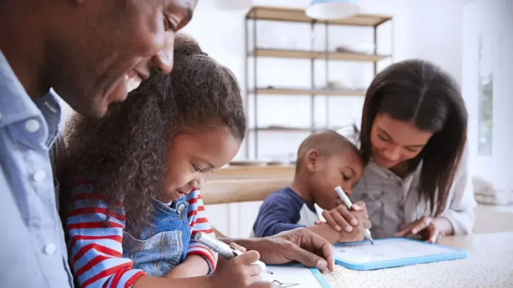 A father, daughter, son, and mother sit at a table as the parents help their children with homework.