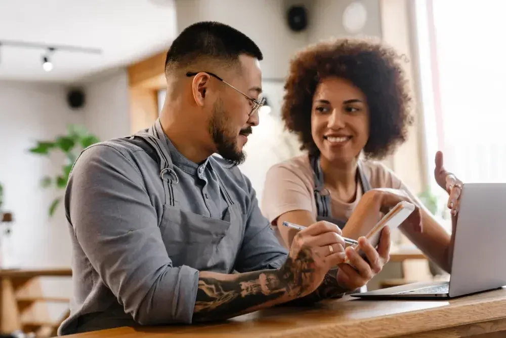 A man and woman who own a business together sit at a counter and go over their BOIR filing checklist. He holds a notepad and pen and an open laptop is in front of her.