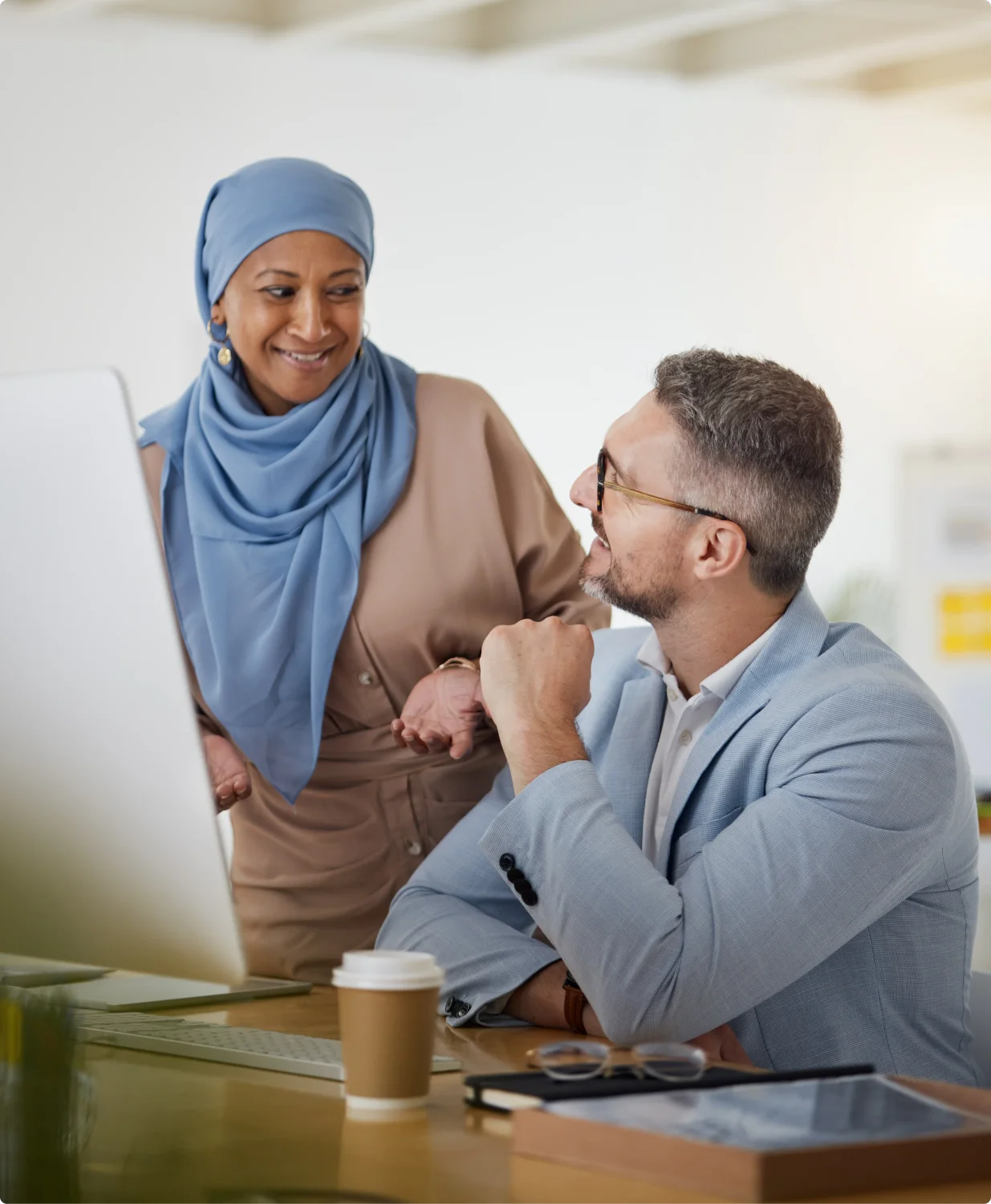 A woman in a blue hijab stands and speaks with a man wearing a business suit and glasses.