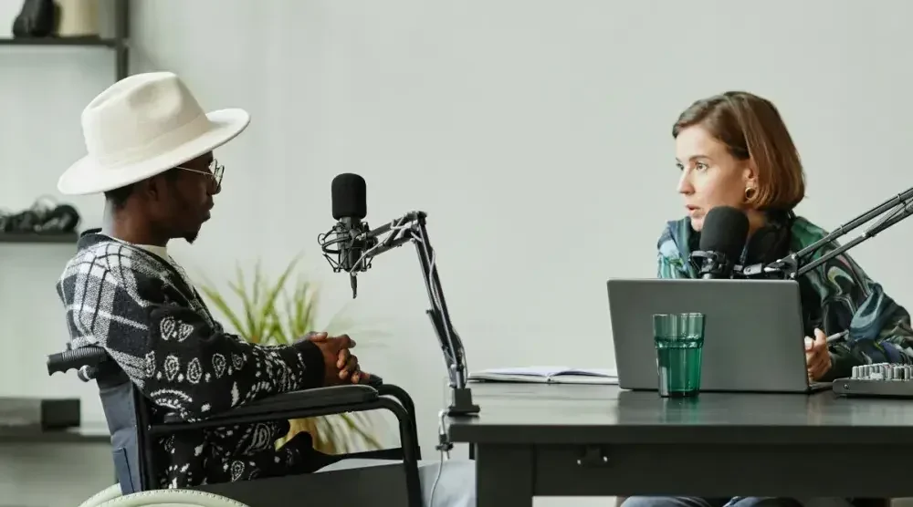 A man wearing a fedora sits in a wheelchair and speaks into a microphone while being interviewed by a podcaster, who sits opposite him behind a desk.