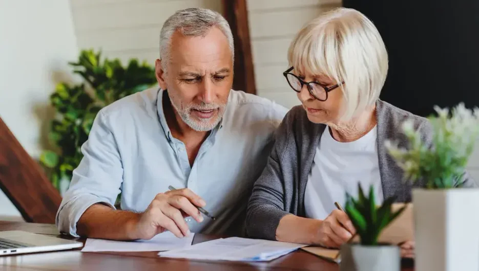Elderly couple sitting closely in cozy living room with lot of green plants, reviewing notes on how to create a living will in simple steps.