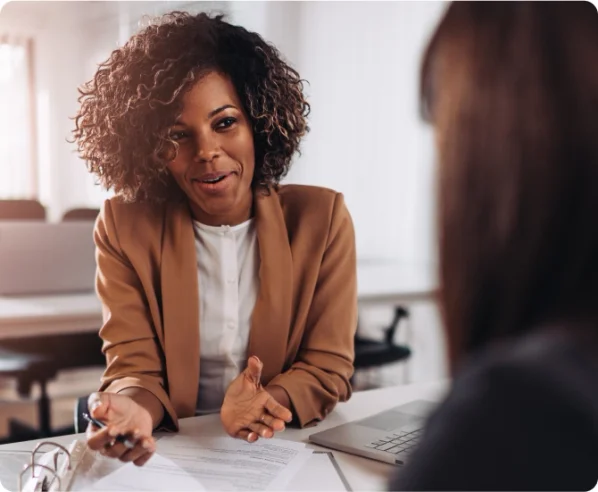 A black woman wearing a brown business suit has a discussion over paperwork with a client.