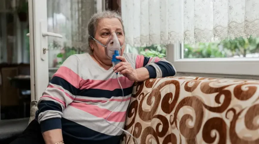 A woman sits on her couch and looks out her large front window while using a portable oxygen mask.