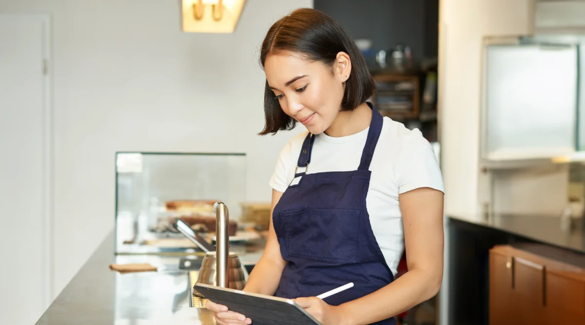 A woman in an apron stands behind the counter of a light-filled cafe and reviews notes on her tablet