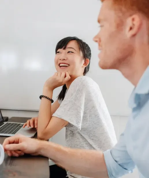 An asian woman in the background sits at her computer and laughs with a man in the foreground.