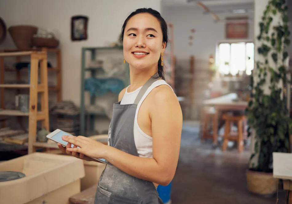 Smiling asian woman wearing a white tank top and a gray apron holding a tablet in a pottery workshop.