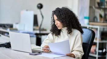 Woman analyzing papers at a desk with a laptop