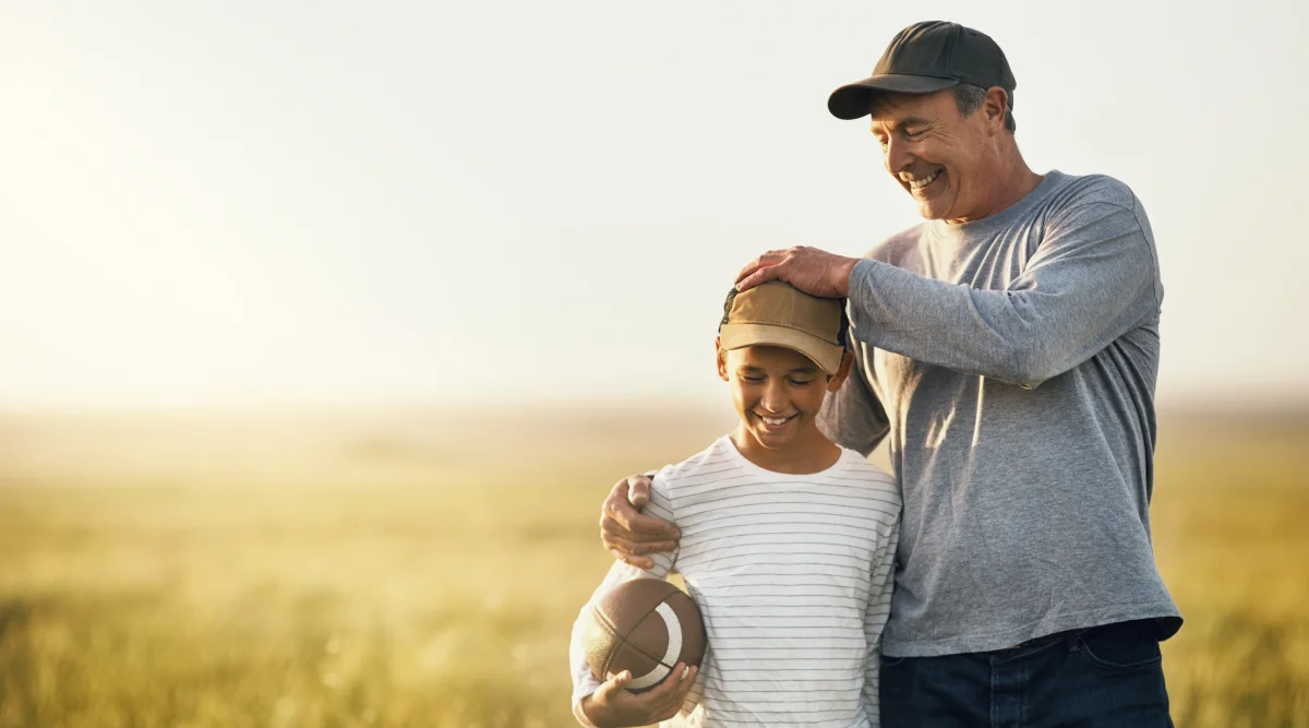 A father wearing a black cap, smiling and holding his son, who has a baseball in his hands, outside on the fields.