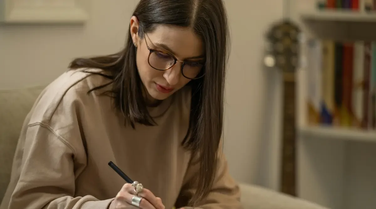A young woman in glasses sits in her office and takes notes in a notebook.