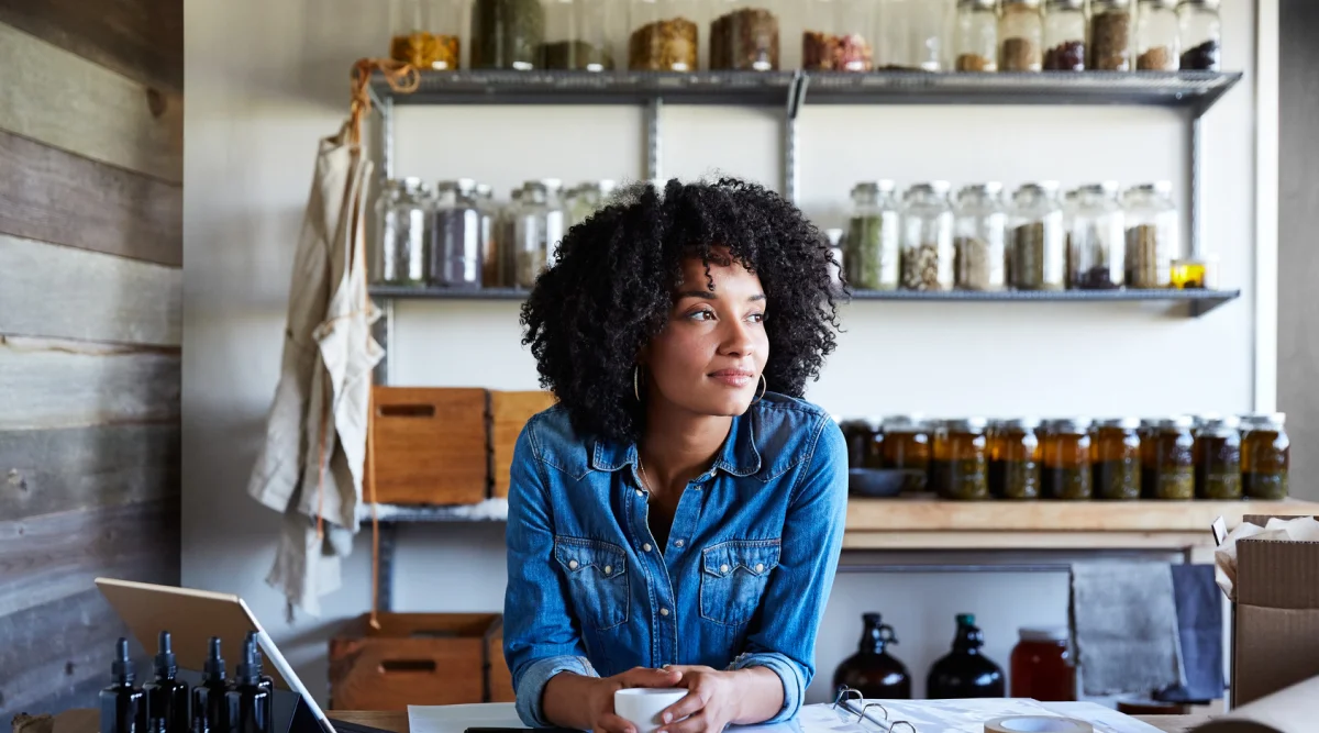 A black woman with natural hair stands behind the counter in her team shop and sips a cup of tea while watching customers browse.