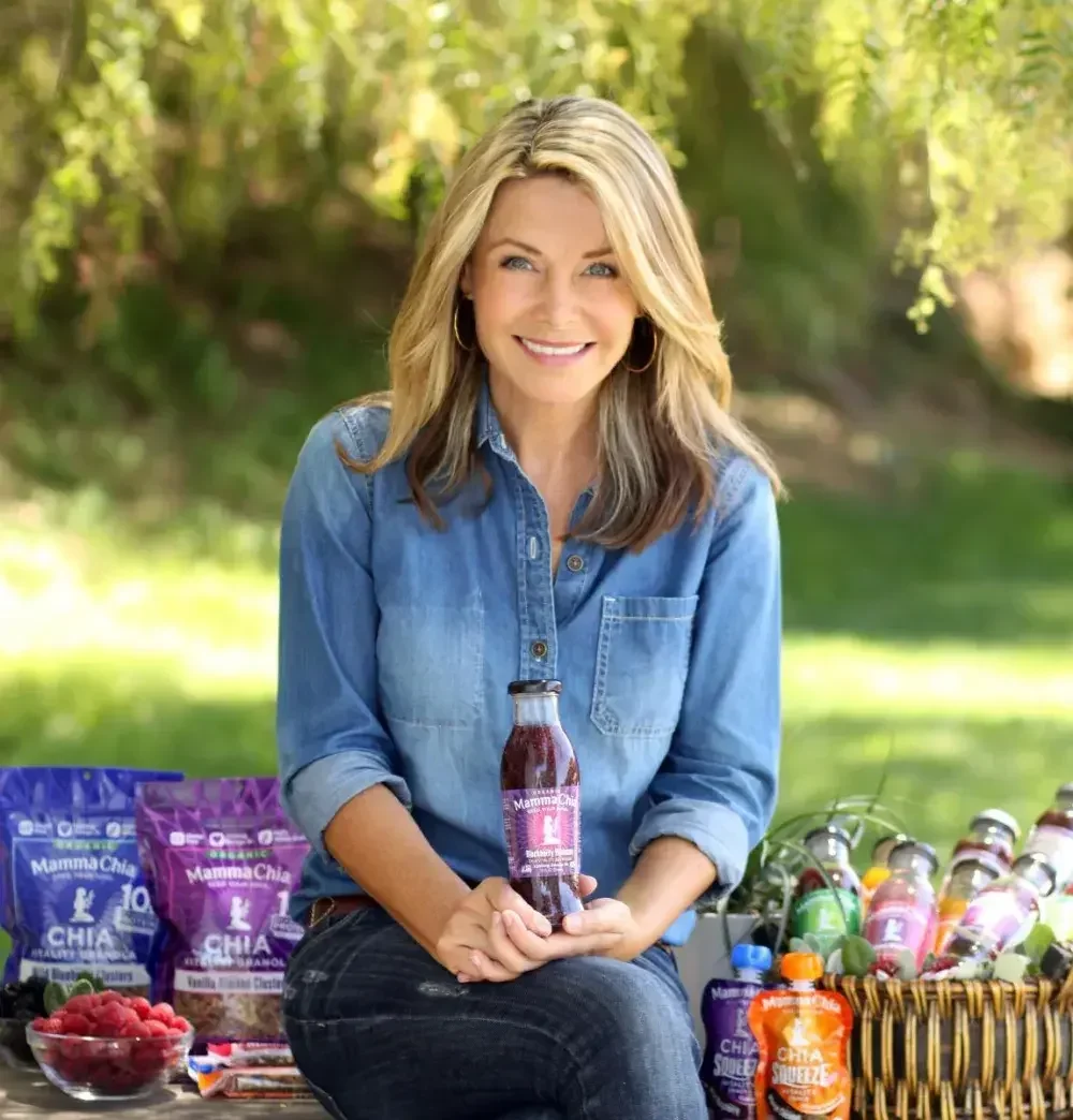 Mamma Chia founder Janie Hoffman sits in a sunny garden surrounded by Mamma Chia products: bags of chia seeds, bottles of chia drink, chia pouches, and fresh fruit.