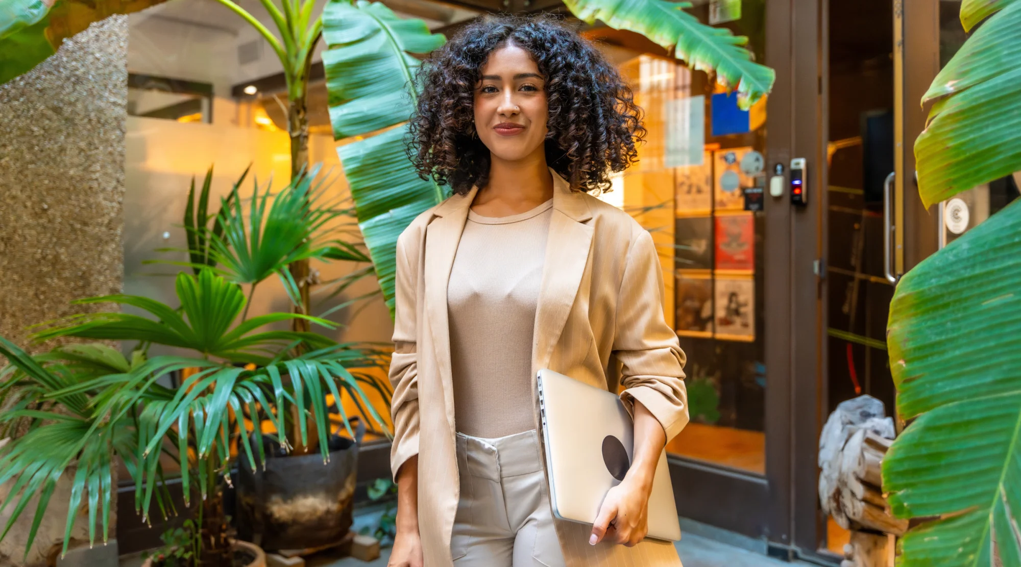 A woman smiles and stands in front of a modern storefront surrounded by tropical greenery.