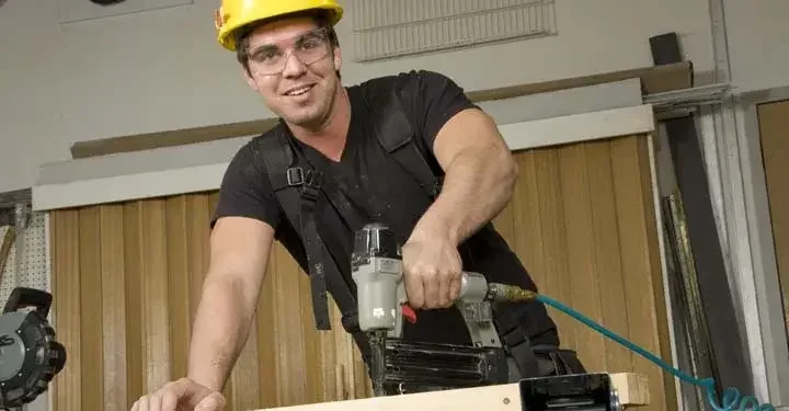 A construction worker in a hard hat and goggles holding a nail gun on a plank of wood.