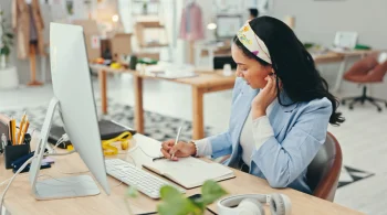 Woman writing in a notebook at a desk with a computer