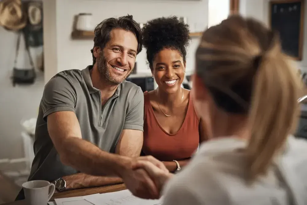A man shakes hands with a estate planning professional seated across a desk from him as his partner, seated next to him looks on and smiles. They have just completed their paperwork for their wills.