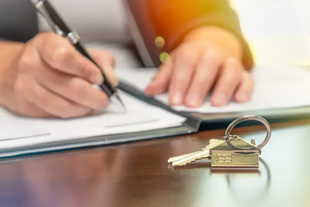 A person signs a quitclaim deed in a folder as she prepares to hand over a set of keys, which are on the desk.