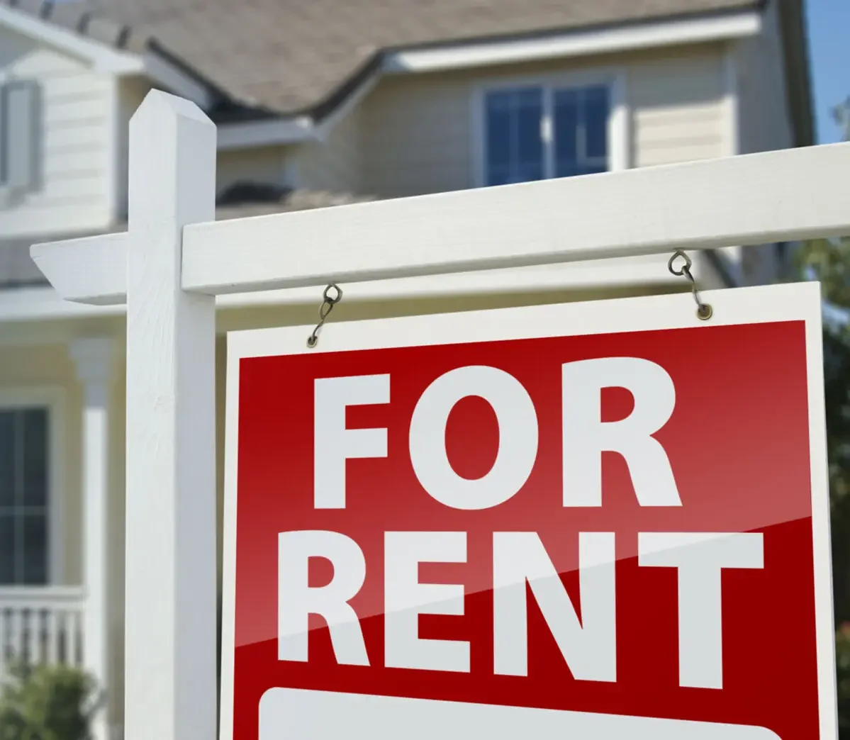 A red and white 'For Rent' sign hanging on a white post in front of a suburban house