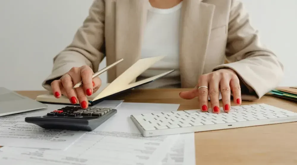 A woman with bright red nails is seated at a desk and working on a calculator with one hand and a computer keyboard with the other hand as she considers bankruptcy options.