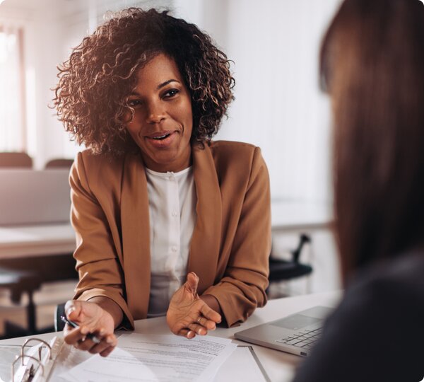 A black woman wearing a brown business suit has a discussion over paperwork with a client.