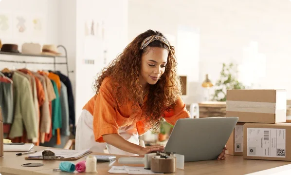 Black woman with her hair pulled back with a headband, wearing a flowy orange blouse and flowy white pants. Looking at her laptop and getting her business license from LegalZoom.