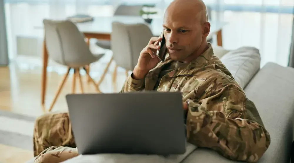 A U.S. service member sits on a couch in uniform, holding a phone to his ear and looking at a laptop screen. 
