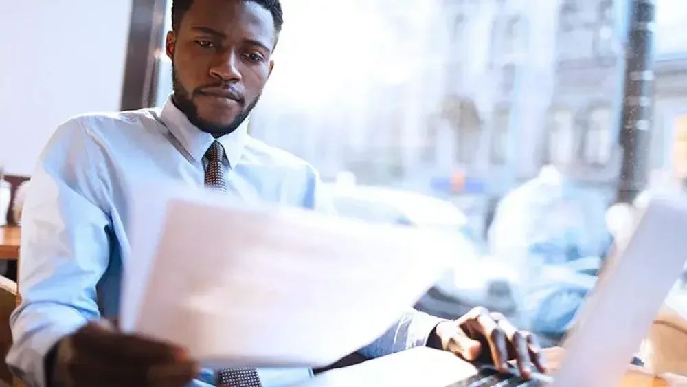 A business owner sits at his desk in his office and reads FinCEN documents he printed out from the laptop computer in front of him.